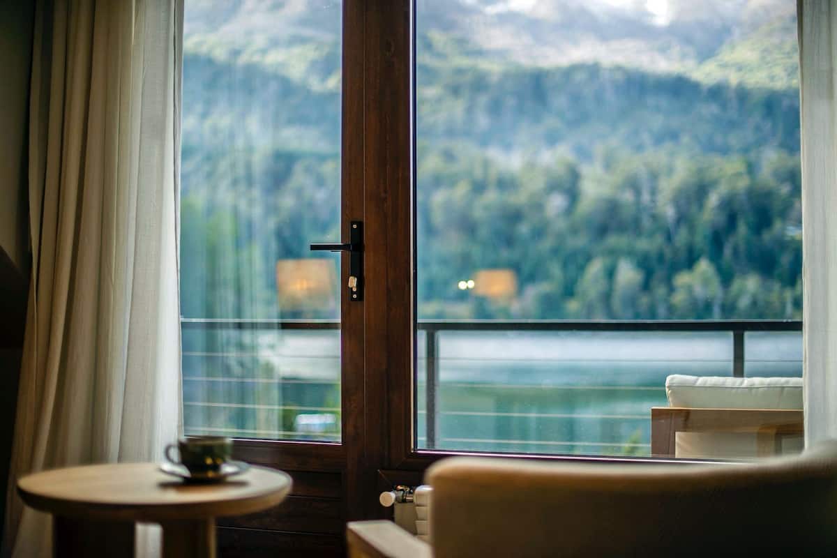 A room with a wooden door, a small round table with a cup, and a view of a forested mountain area.