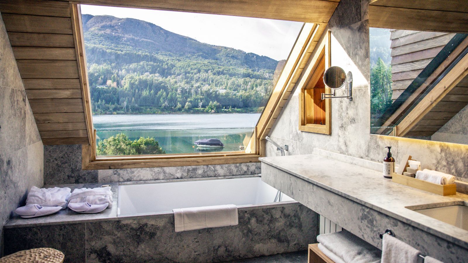 Un baño con bañera, lavamanos, espejo y vistas a un lago y un bosque.