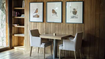 Pared de madera con tres cuadros de pájaros enmarcados, una mesa de madera con dos sillas y una estantería con libros y adornos.