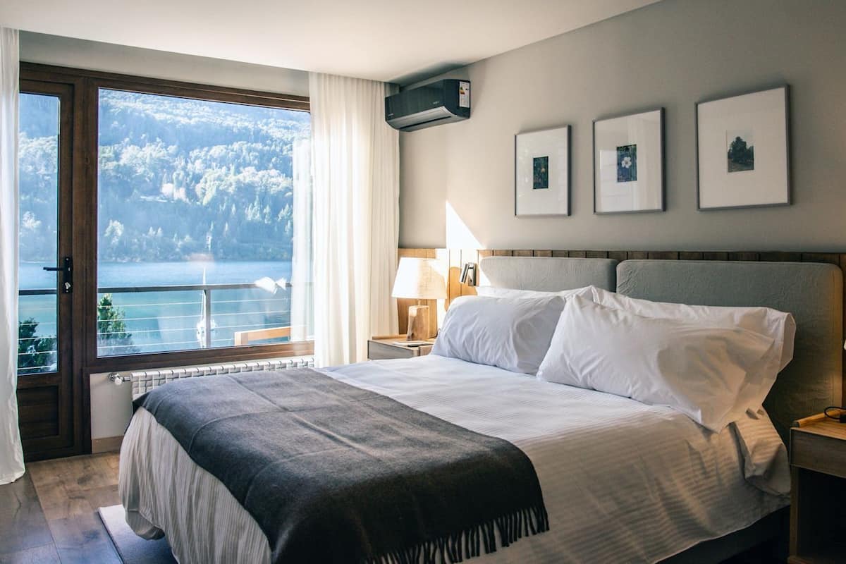 A bedroom with a large bed, a nightstand with a lamp, a window with a view of a mountain and lake, and three framed pictures on the wall.