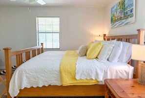 Ein Schlafzimmer mit einem hölzernen Bett, Bettwäsche in Weiß und Gelb, einem Fenster mit Jalousien, einem Gemälde an der Wand und einer Lampe auf einem Nachttisch.