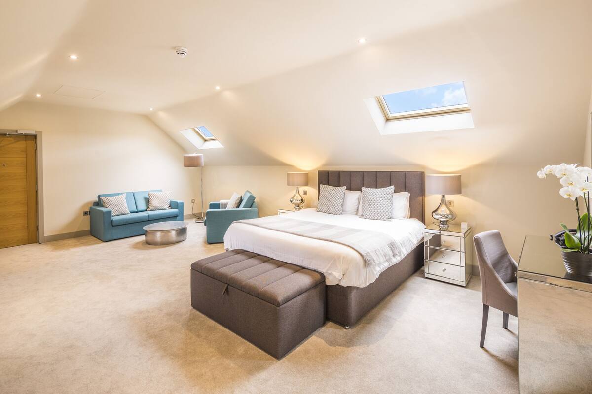 A bedroom with a large bed, a sofa, a desk, and a small table. There are skylights on the ceiling.