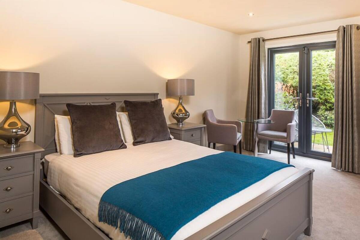 A hotel room with a bed, two bedside tables, a chair, a small table, and a balcony with curtains.