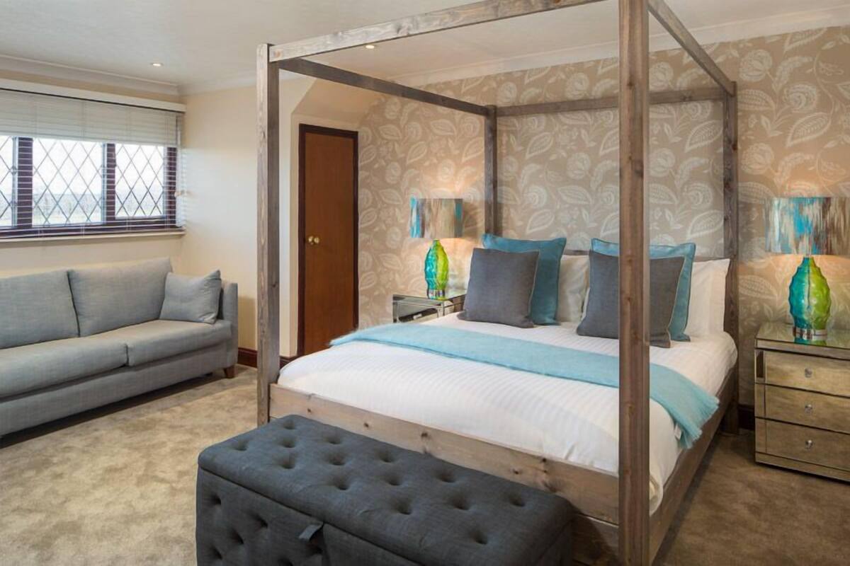 A four-poster bed with a wooden frame, a grey upholstered bench, and a grey sofa in a bedroom with patterned wallpaper and a window with blinds.