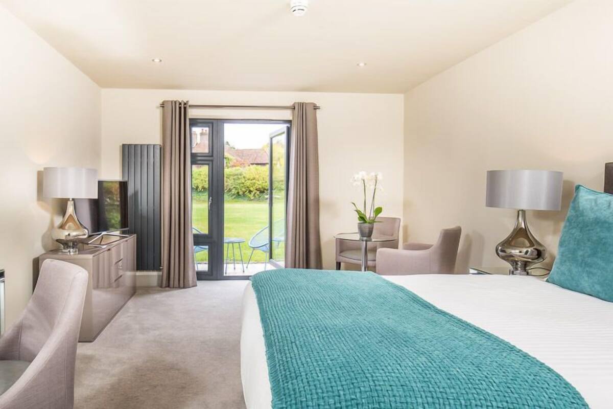 A hotel room with a large bed, two bedside tables with lamps, a chair, a small table with a plant, and a view of a garden through a sliding door.
