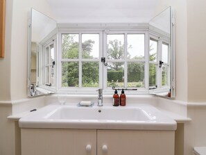 A bathroom with a large white sink, a mirror, and a window with a view of trees. - Magdalen Cottage (Banbury)