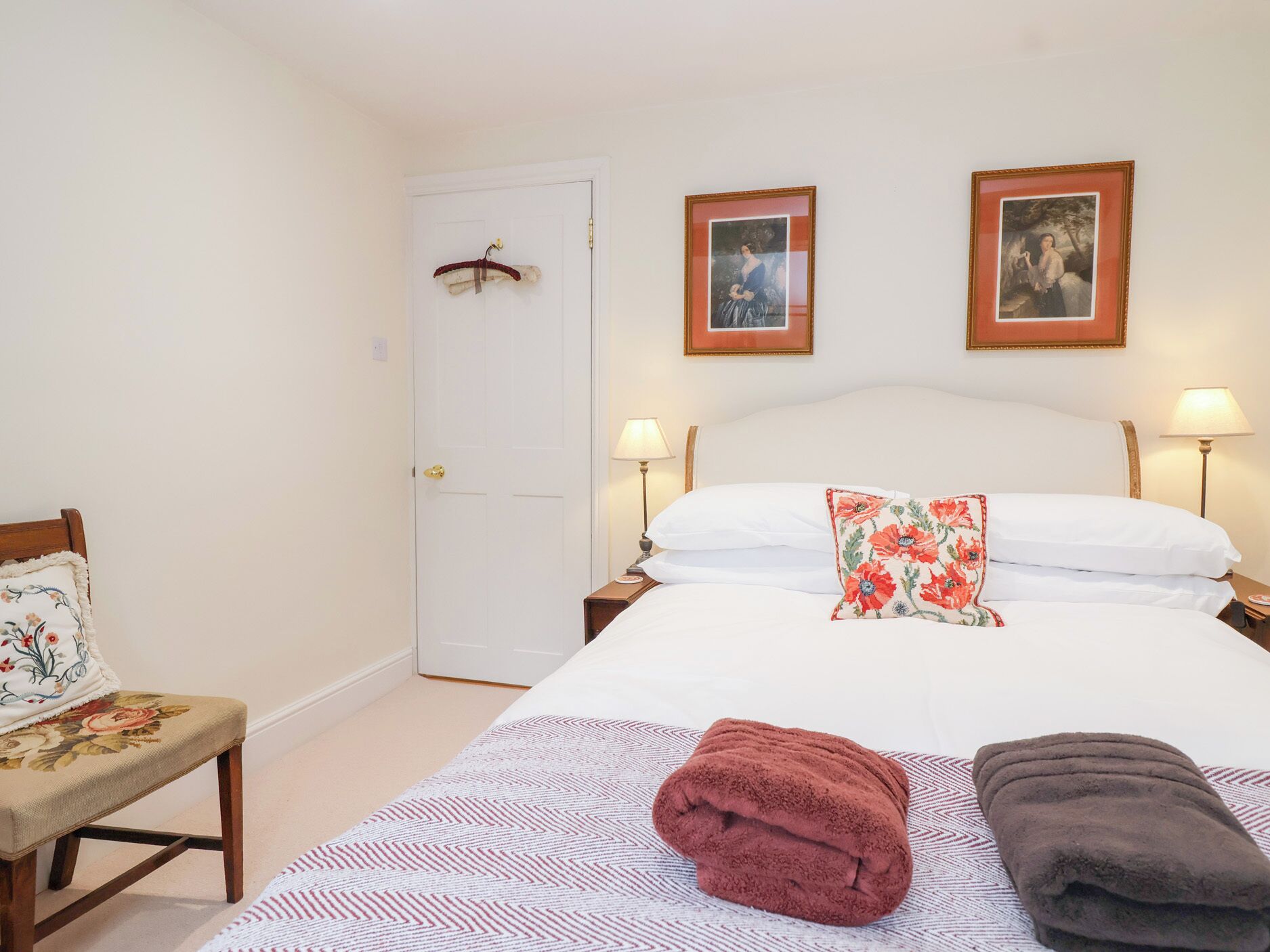 A neatly arranged bedroom with a bed, two bedside lamps, a door with a coat hanging, and a chair with a cushion.