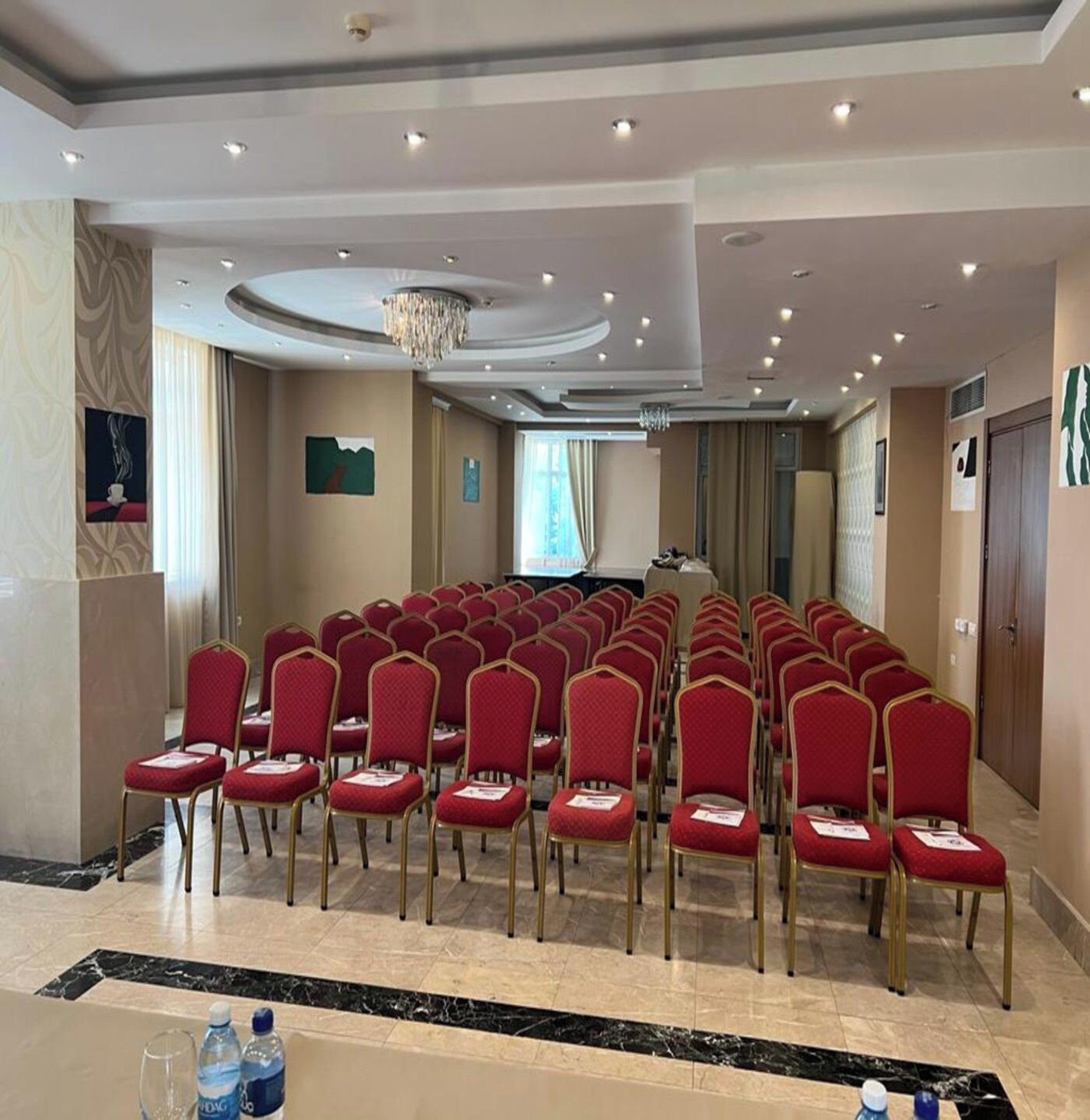 Un salón de conferencias con hileras de sillas rojas, una araña y una pantalla de proyector.