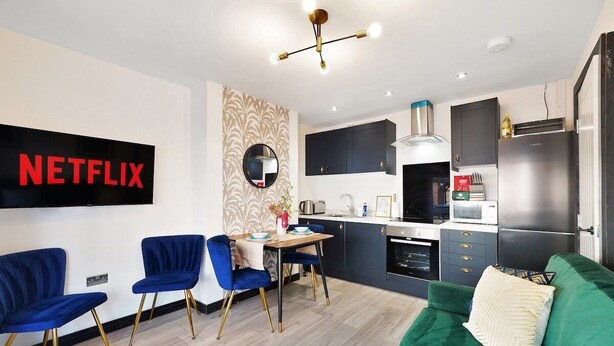 Ein modernes Wohnzimmer mit einem grünen Sofa, einem Essbereich mit blauen Stühlen, einer Küche mit dunklen Schränken und einem an der Wand montierten Fernseher, der "NETFLIX" anzeigt.