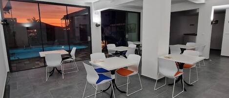 Un comedor moderno con mesas y sillas blancas, amplias ventanas que ofrecen vista a una piscina y al atardecer.
