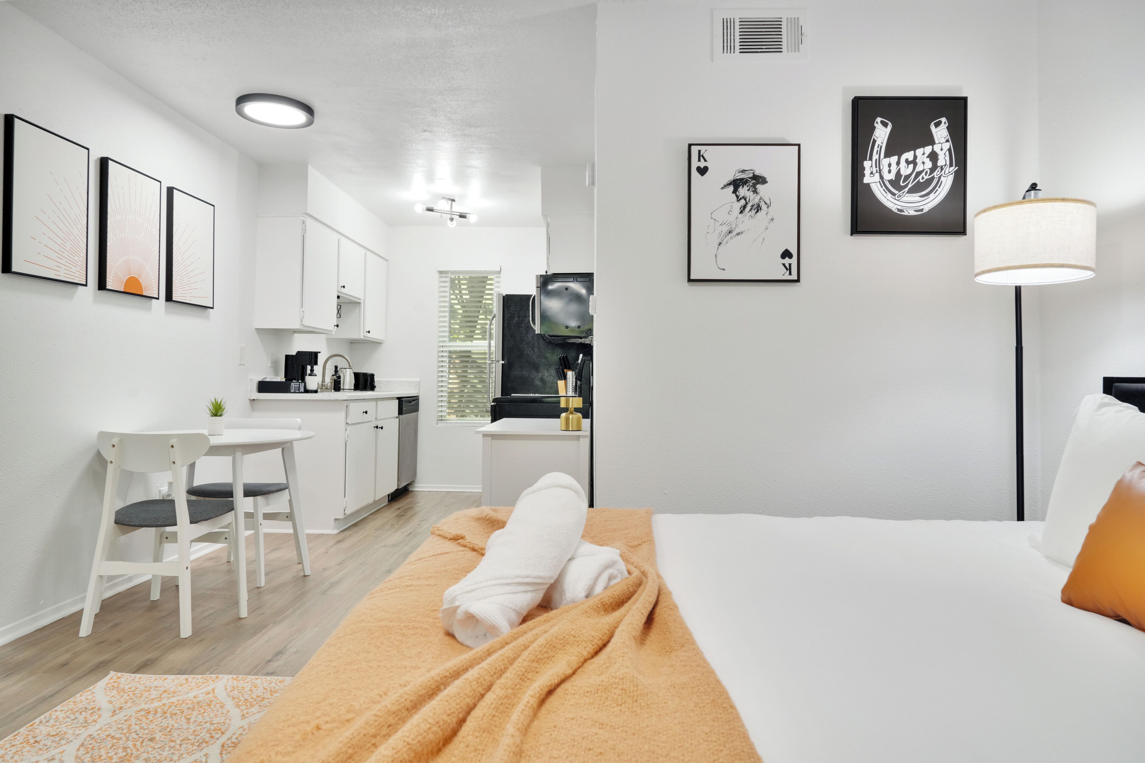 A modern hotel room with a bed, a desk, a chair, and a kitchenette.