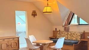 A cozy, well-lit room with a wooden dining table, two white chairs, a floral sofa, and a window with curtains. - In & Out boarding house comfort for four XL rooms (Stolberg)