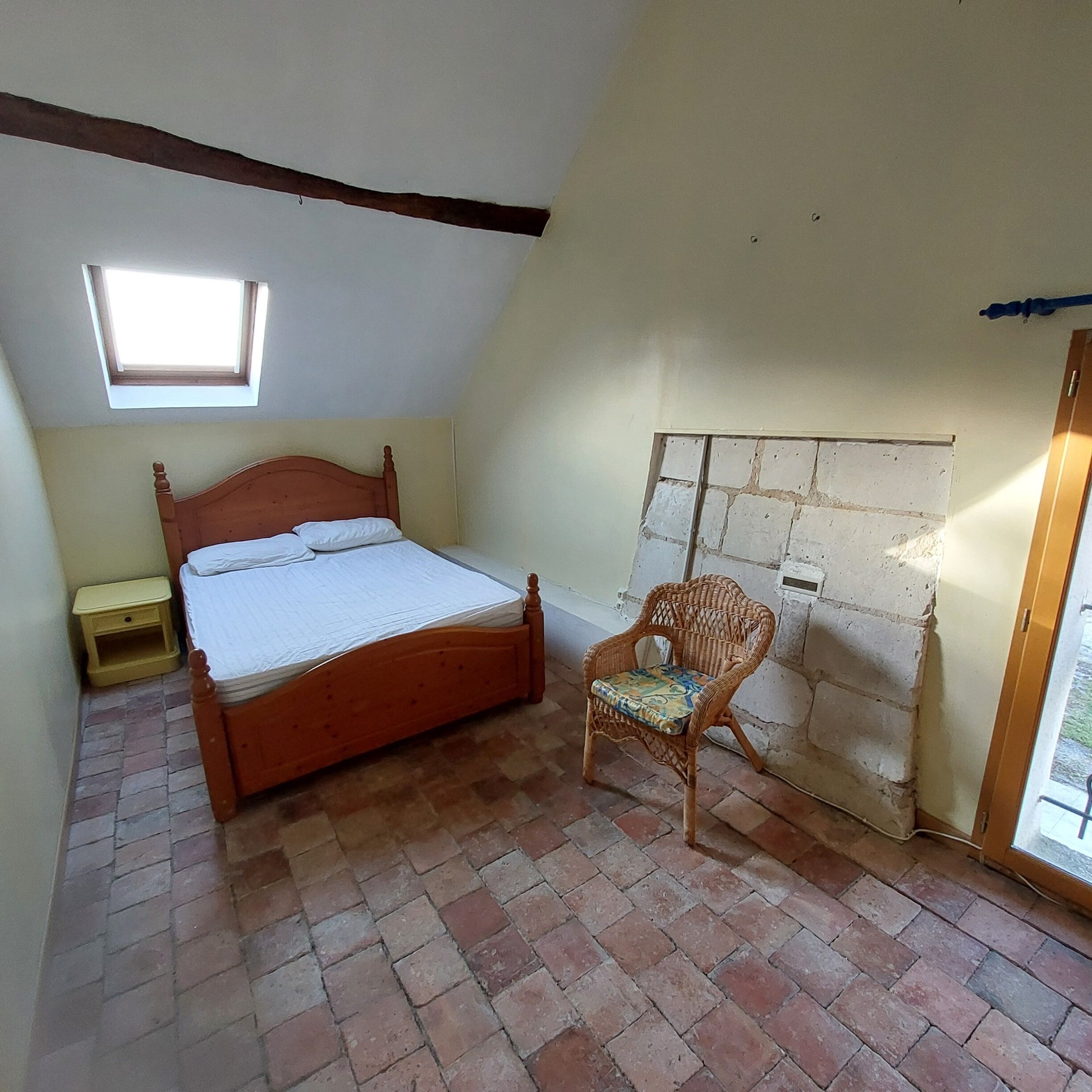 Un dormitorio con cama de madera, una mesita de noche pequeña, una silla de mimbre, techo inclinado y suelo de ladrillo.