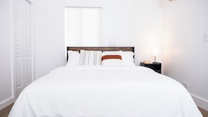 A neatly made bed with white bedding and patterned pillows, a bedside table with a lamp, and a window with blinds.