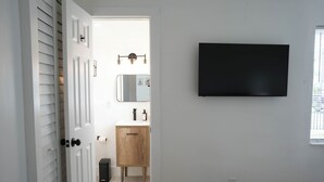 A modern bathroom with a white door, a wooden cabinet, a mirror, and a black TV mounted on the wall. - Comfort Living 2BR Deluxe Suite (West Palm Beach)