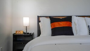 A neatly made bed with white bedding, a black and orange pillow, and a bedside table with a lamp and a digital clock displaying 10:25. - Comfort Living 2BR Deluxe Suite (West Palm Beach)