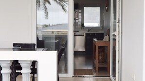Una cocina moderna con electrodomésticos blancos, un comedor con mesa de vidrio y sillas de mimbre, y un ventanal corredero que da a un balcón con vistas a palmeras.