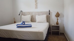 Una cama bien hecha con sábanas blancas y una toalla azul y blanca a rayas, una mesita de noche con un florero, y una lámpara fijada a la pared.