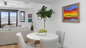 A modern living room with a round dining table, grey sofas, and a large window overlooking a scenic view. - Hummingbird House — Private Rooftop Terrace, Panoramic Ocean & Pier Views, 2BR/2BA Luxury Home · Steps to Avila Beach · Fireplace · Gourmet Kitchen · Beach Gear & Bikes Included · Sleeps 5 · Managed by Solmaré Stays (Avila Beach)