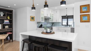 A modern kitchen with white cabinets, a central island with bar stools, pendant lighting, and a bookshelf with books and decorative items. - Hummingbird House — Private Rooftop Terrace, Panoramic Ocean & Pier Views, 2BR/2BA Luxury Home · Steps to Avila Beach · Fireplace · Gourmet Kitchen · Beach Gear & Bikes Included · Sleeps 5 · Managed by Solmaré Stays (Avila Beach)