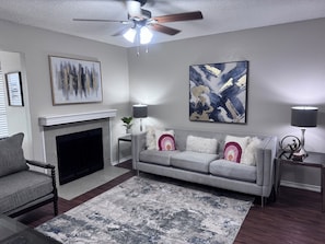 Un salon avec une cheminée, un canapé gris, une table basse et un ventilateur de plafond.