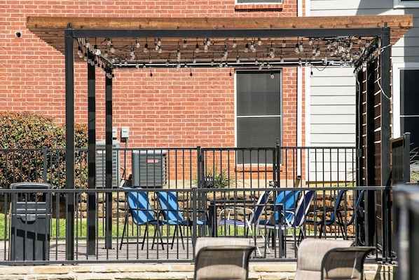 Un espace patio agrémenté d’un mur en briques, d’une pergola illuminée de guirlandes lumineuses et de plusieurs chaises.