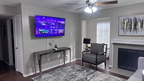 A living room with a television, a fireplace, a ceiling fan, and a decorative painting. - I012 - Upgraded 2BD | Pool | DFW South 183/161 (Irving)