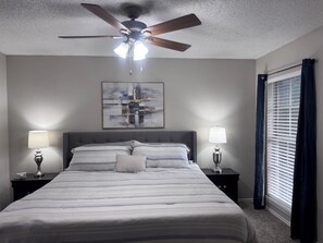 A hotel room with a bed, two bedside lamps, a ceiling fan, a window with blinds, and a wall-mounted abstract painting. - I012 - Upgraded 2BD | Pool | DFW South 183/161 (Irving)