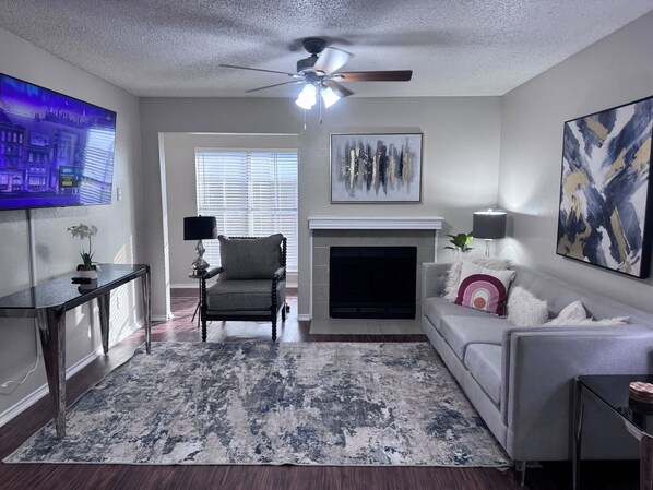 A living room with a fireplace, a television, and a ceiling fan. - I012 - Upgraded 2BD | Pool | DFW South 183/161 (Irving)