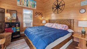 A wooden-paneled bedroom with a large bed, a wall clock, a compass, and a painting of surfboards. - The Barn: Private Pool | Hot Tub (Kill Devil Hills)