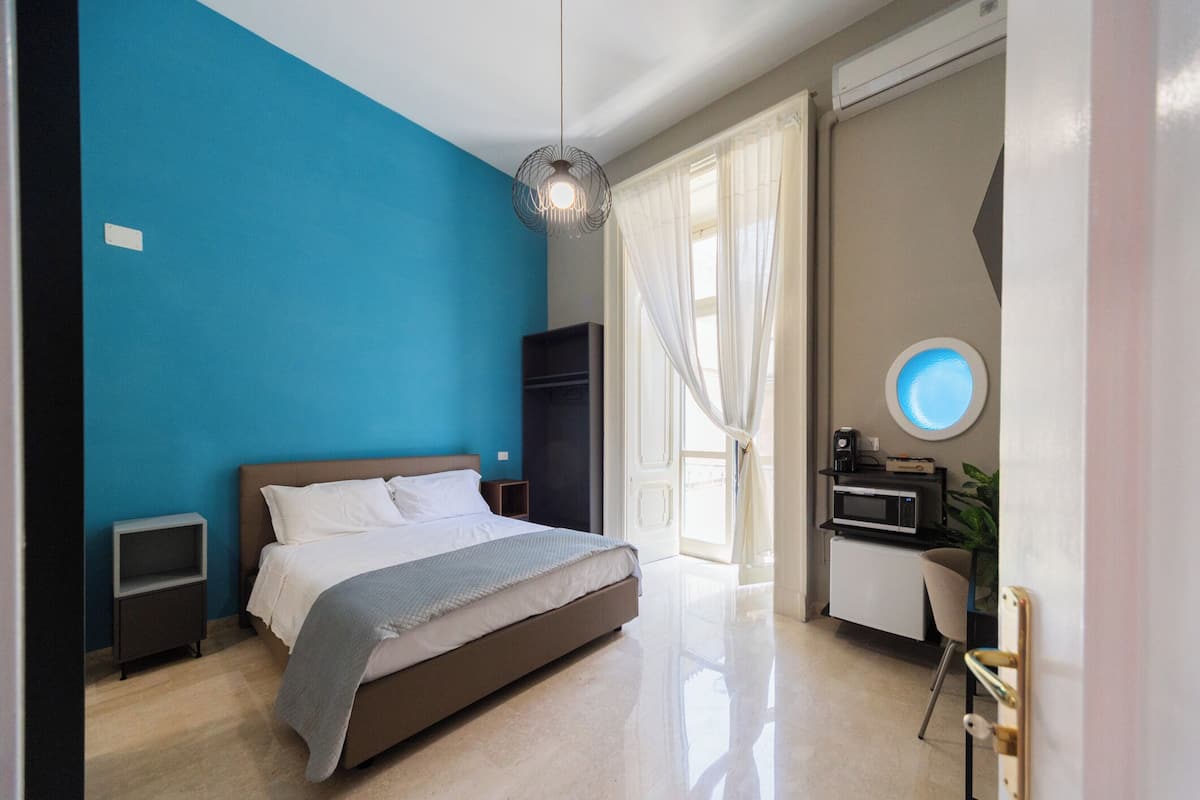 Un dormitorio moderno con cama, mesitas de noche, un armario, un escritorio con silla, un espejo redondo y una ventana con cortinas.