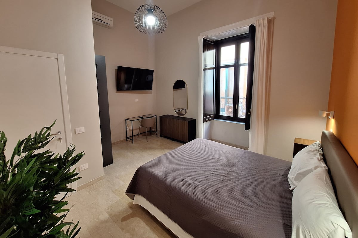 Un dormitorio con cama, escritorio, silla, televisor, ventana con cortinas y una planta.