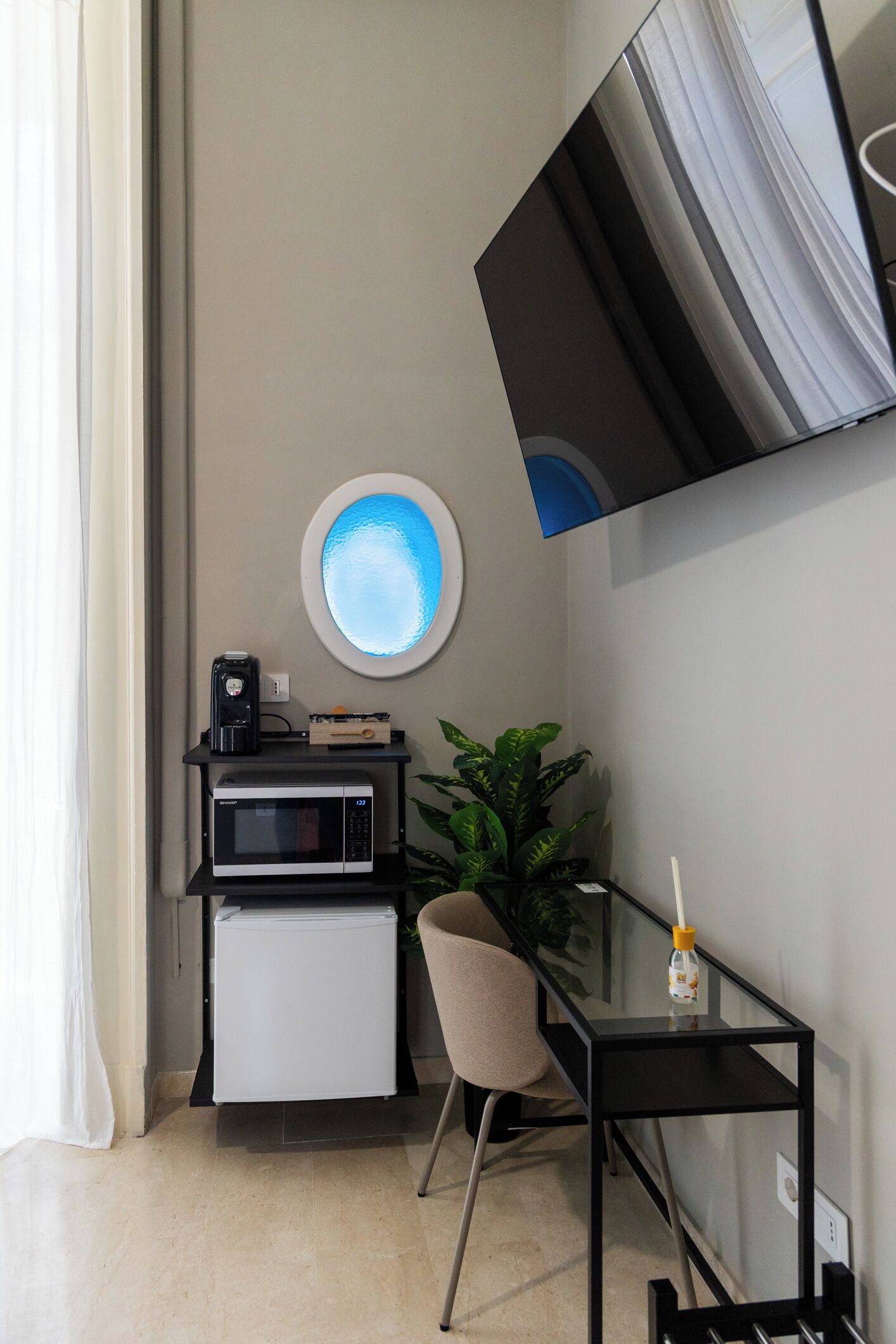 A compact living space with a kitchenette, a flat-screen TV mounted on the wall, a small refrigerator, a microwave, a coffee maker, a potted plant, and a glass table.