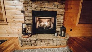 A stone fireplace with a log fire, surrounded by wooden walls and a rug on the floor. - Peaceful Cabin: Trails, Winery, Fire-pit & 12 Acr (Newland)
