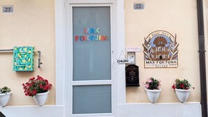 A building facade with a sign reading "MAX FORTUNA" and another sign for "MAX FORTUNA ITALIAN GUEST HOUSE RAGUSA SICILY". There are three potted plants on the ground level, a mailbox, and a decorative plaque on the wall. - MAX-FORTUNA (Ragusa)