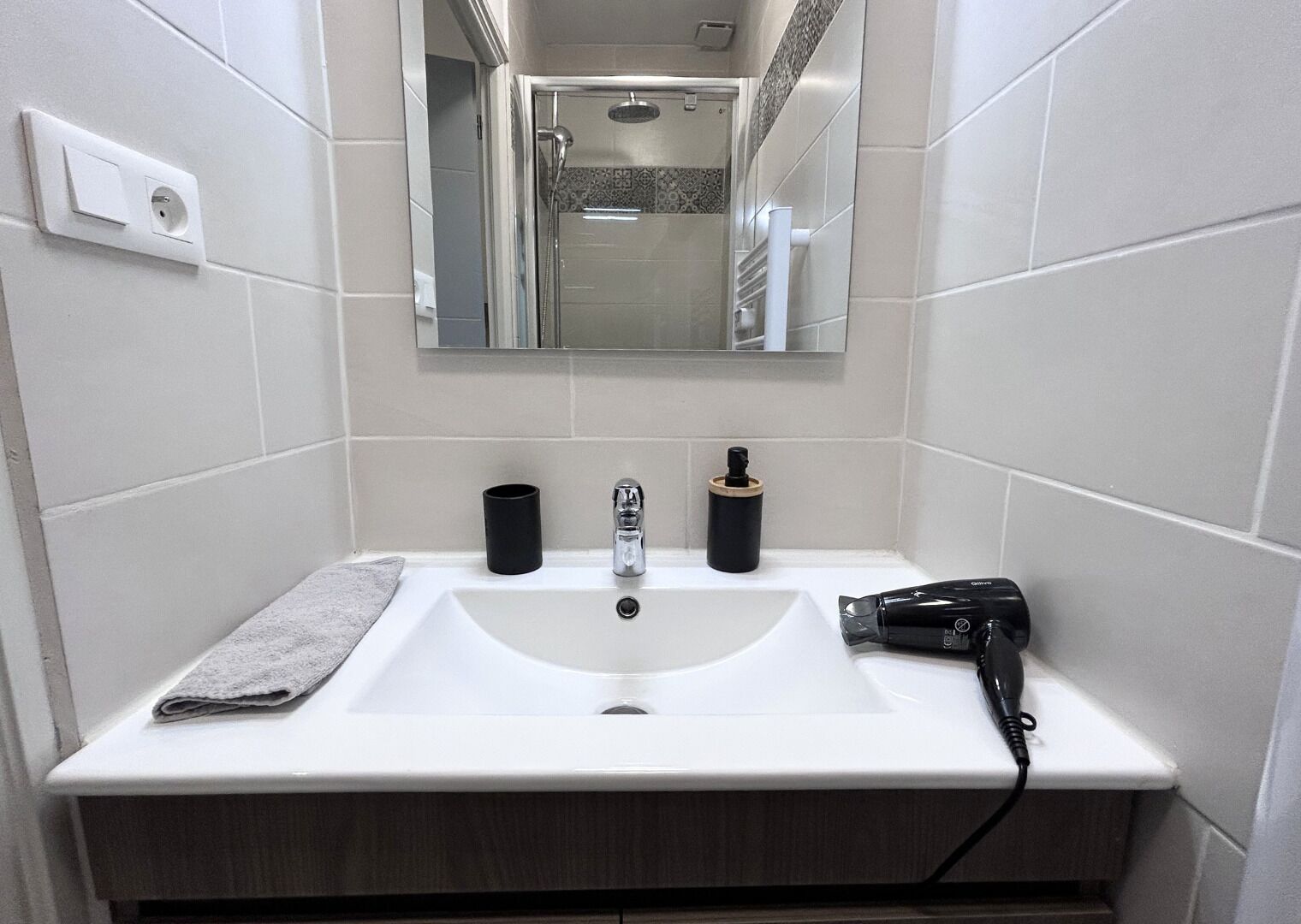 Un baño con un lavamanos blanco, un espejo encima y una ducha a la vista al fondo.