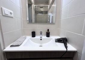 Un baño con un lavamanos blanco, un espejo encima y una ducha a la vista al fondo.