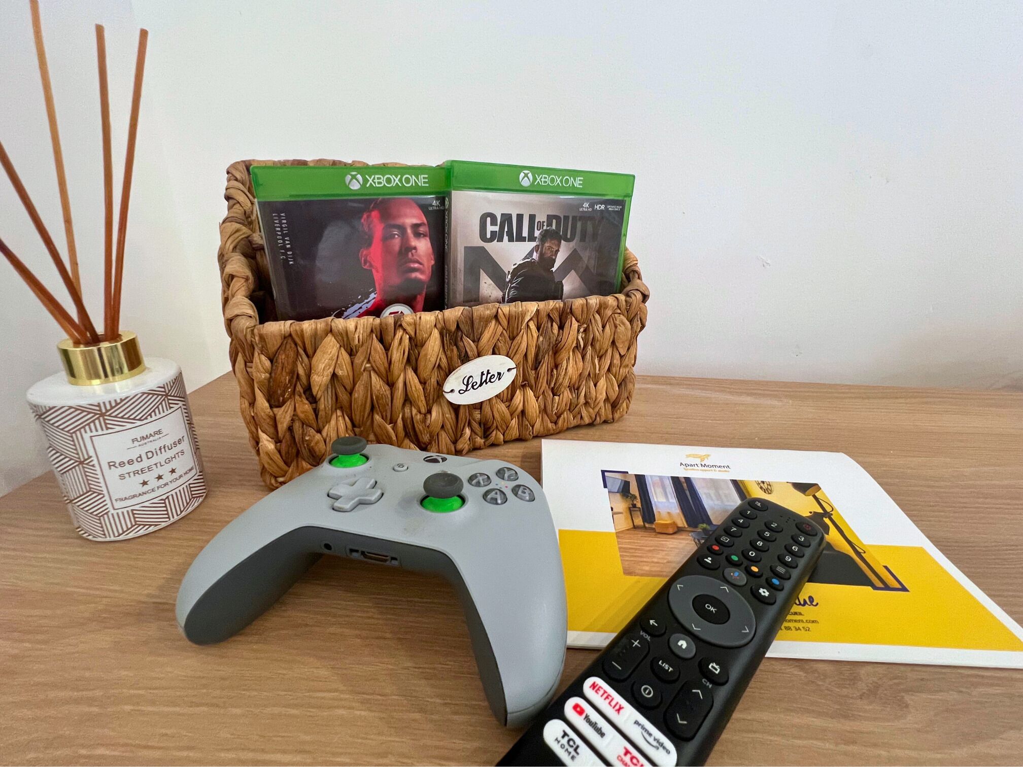 Un equipo de juegos con un control de PlayStation, dos juegos de Xbox One, un control remoto y una revista sobre una mesa de madera.