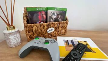 Un equipo de juegos con un control de PlayStation, dos juegos de Xbox One, un control remoto y una revista sobre una mesa de madera.