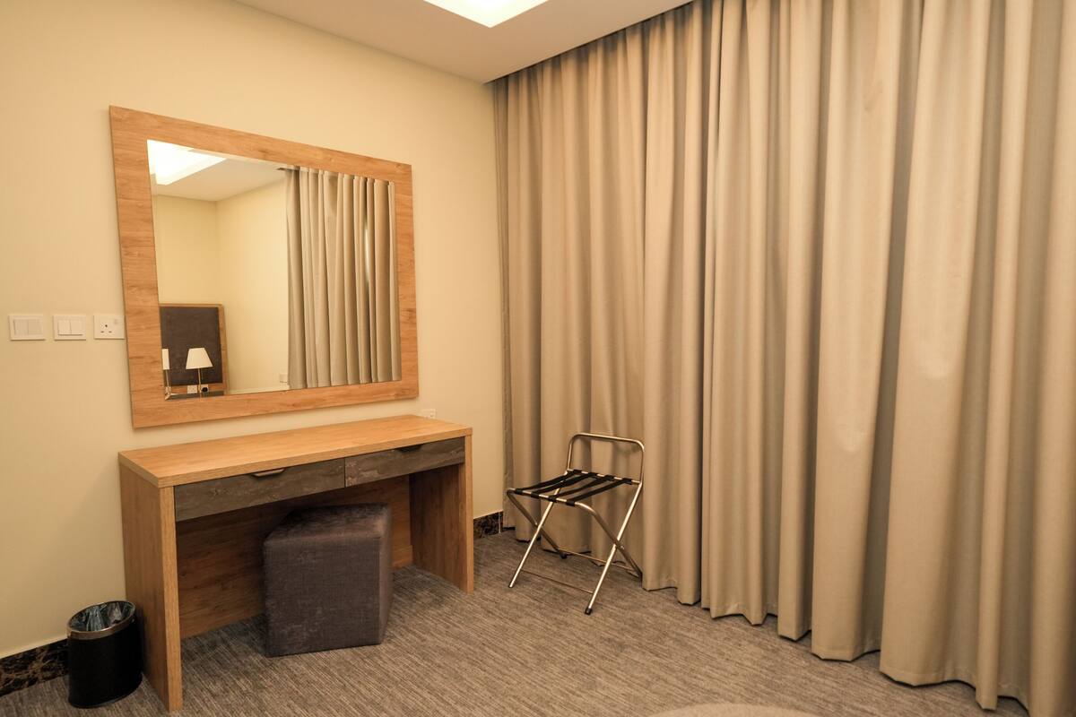 A hotel room with a wooden desk, a mirror, a chair, and beige curtains.