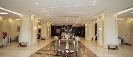 A spacious hotel lobby with a central round table, seating arrangements, and a reception desk.