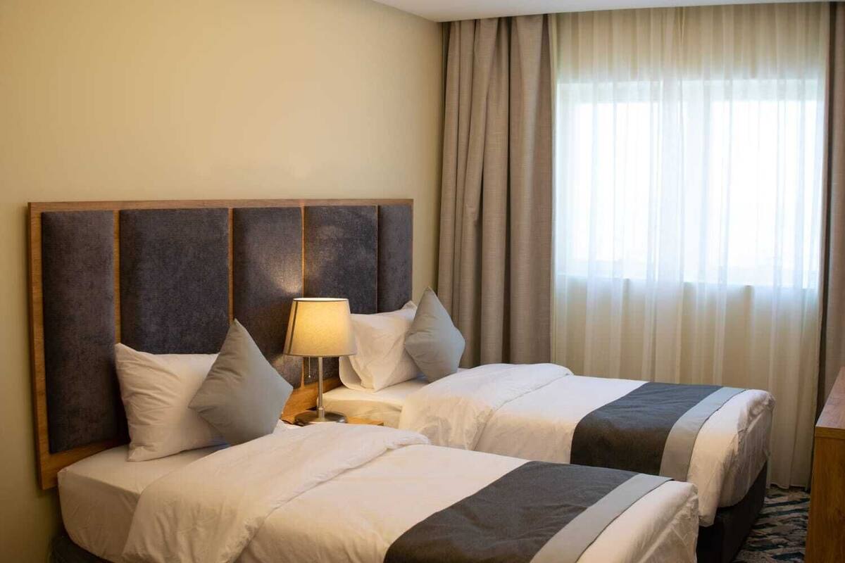 A hotel room with two beds, a nightstand with a lamp, and a large window with curtains.