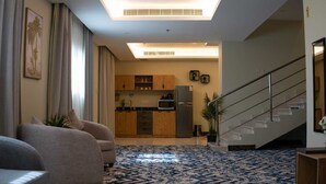 A modern living room with a patterned carpet, a sofa, a dining area with wooden cabinets, and a staircase. - benzrthotel (Al-Hofuf)
