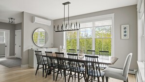 A dining area with a large table, multiple chairs, a chandelier, and a view through large windows. - Downtown Coastal Charm Cottage in Kennebunk (Kennebunk)
