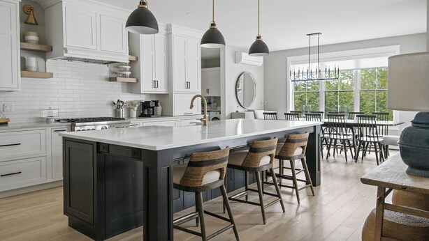 A modern kitchen with white cabinets, a large island with bar stools, pendant lighting, and a view of the outdoors through large windows. - Downtown Coastal Charm Cottage in Kennebunk (Kennebunk)
