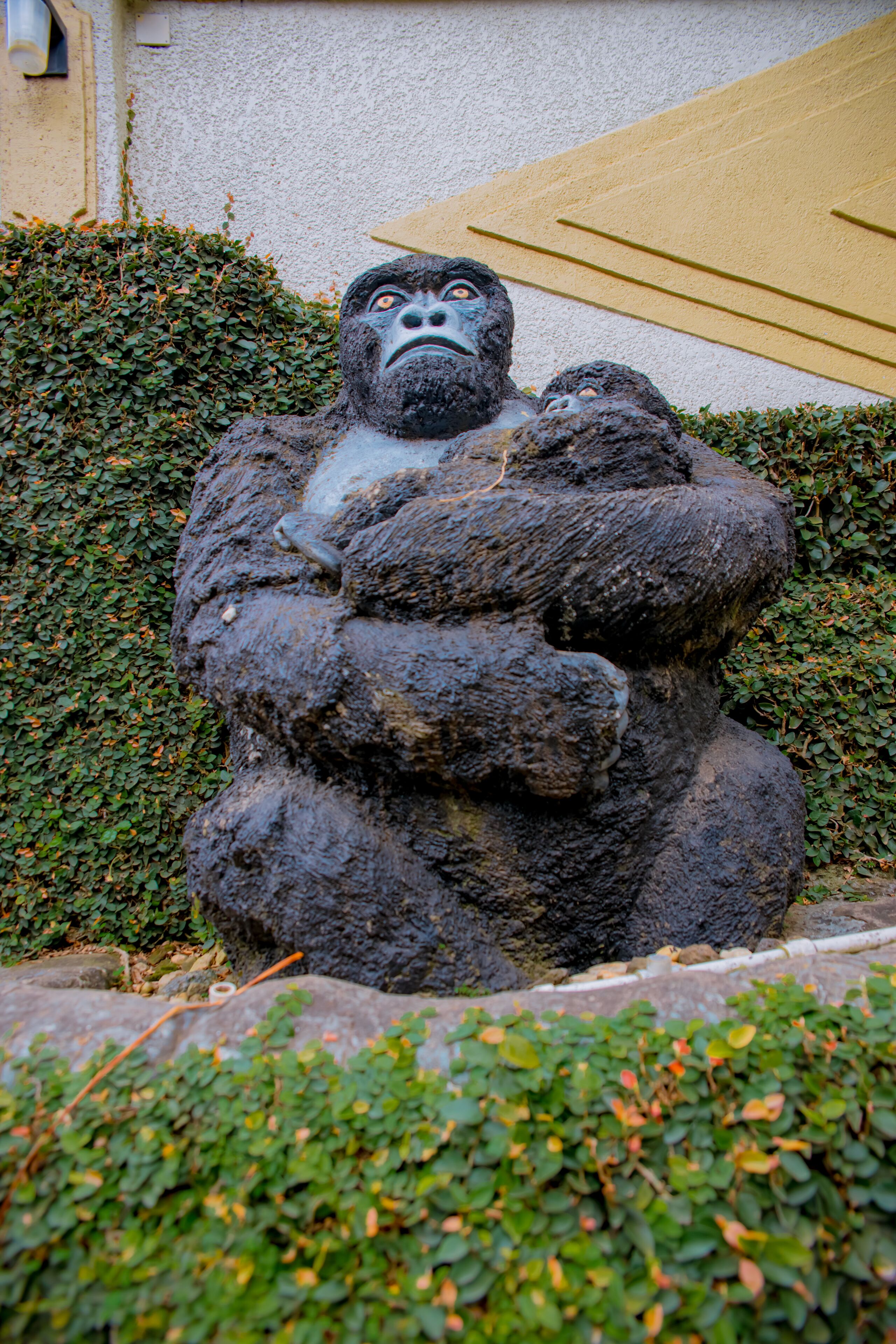 A large gorilla statue with a baby gorilla, surrounded by greenery and a building in the background.