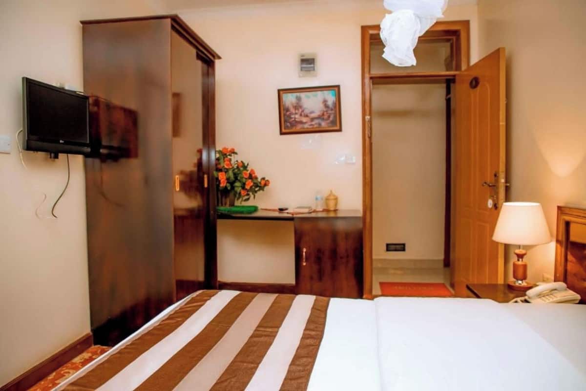 A hotel room with a bed, a desk, a television, a painting, a lamp, and a telephone.