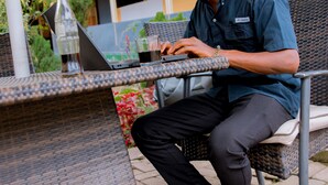 A man sitting at an outdoor wicker table with a laptop, a bottle, and a glass. - INEZA HOTEL (Ruhengeri)