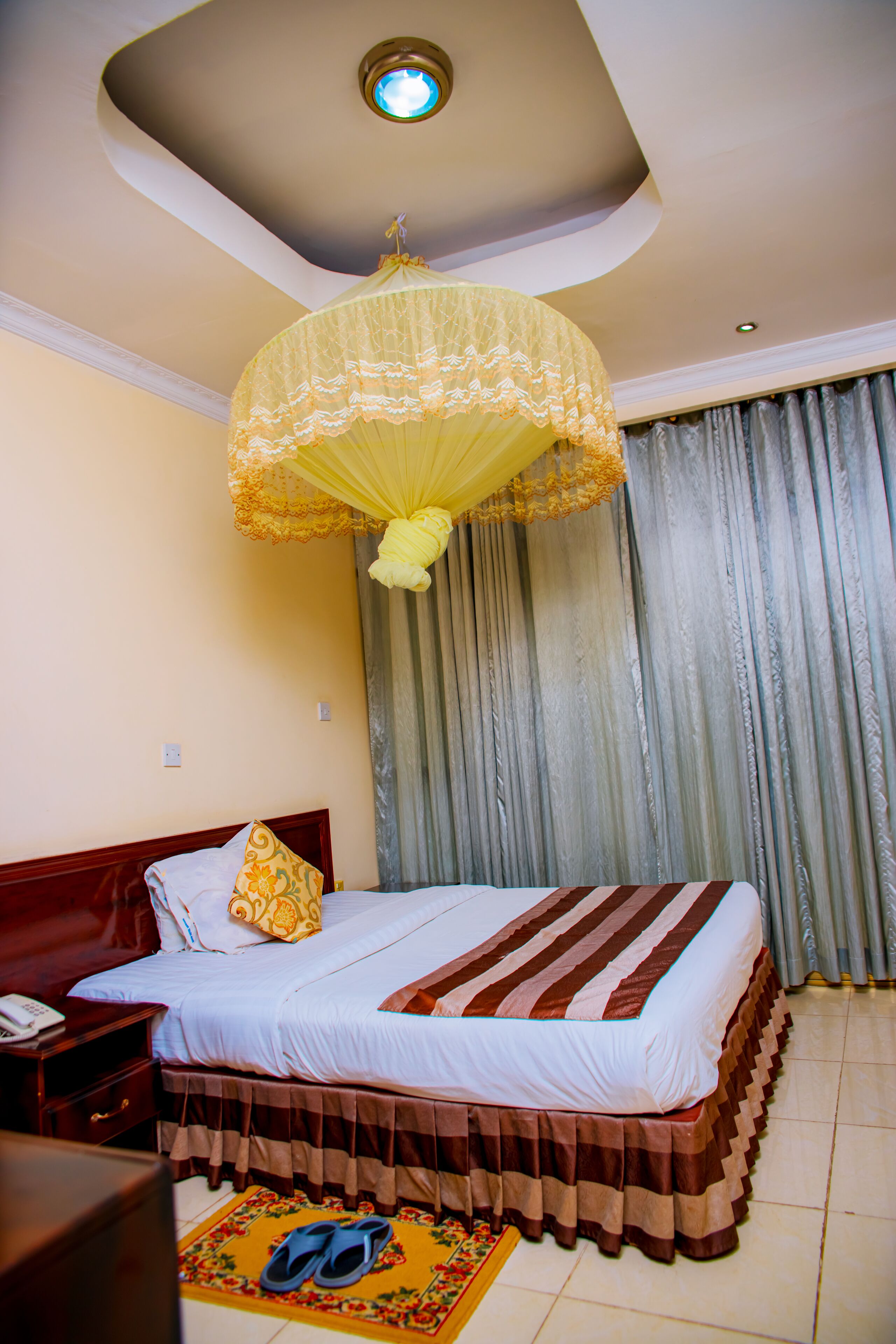 A neatly made bed with a striped bedspread, a bedside table with a phone, and a pair of slippers on a mat.