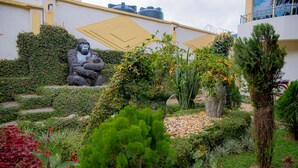 A landscaped garden with a gorilla statue, various plants, and a building with yellow accents and water tanks. - INEZA HOTEL (Ruhengeri)